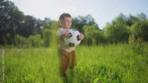 Young boy is holding soccer ball. Male child holds a soccer ball outdoors. A casual field of greenery and grassy nature in summer. A young boy is grasping a lifestyle soccer ball.