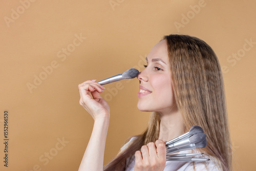 A beautiful girl with big eyes smiles and holds makeup brushes in her hands on a brown background.