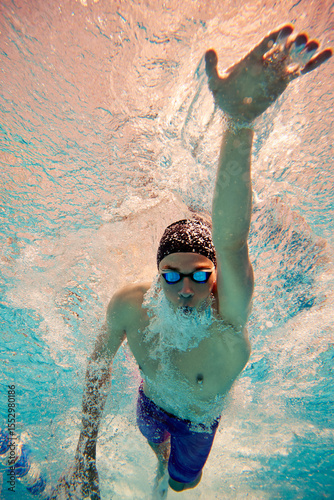 An underwater perspective shows a male swimmer in his 20s, performing a freestyle stroke in a brightly lit pool. Concept of competitive swimming and athletic endurance.