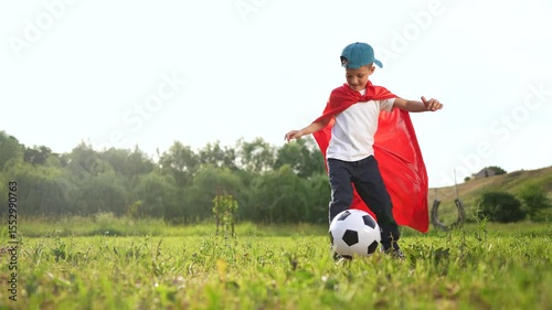 Young boy in red cape playing with soccer ball. Boy is playing soccer outdoors. Child is kicking a ball in the grass while being energetic. A young boy wearing a red cape is kicking a lifestyle.