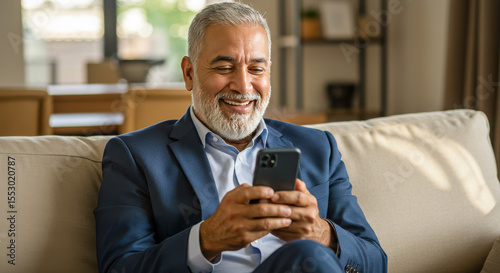 Smiling happy senior mature middle aged man holding cell mobile phone using smartphone sitting at home on couch, scrolling social media, checking financial apps, buying online, texting messages.