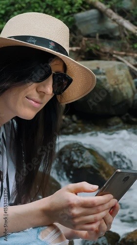 Portrait of curly girl in forest looking in smartphone hiking near river. Young woman hiker in mountain hike finding route on map in phone in Summer. Tourism, travel, hike, trip, adventure concept.