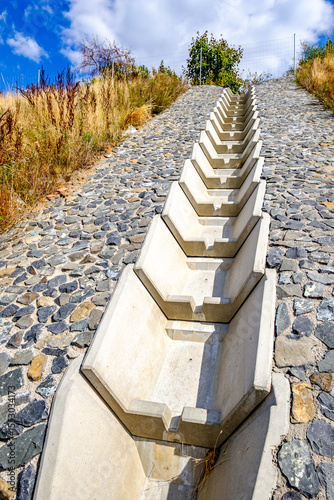 Concrete drainage channel on a hillside lined with stones surrounded by dry grass and shrubs under a blue sky