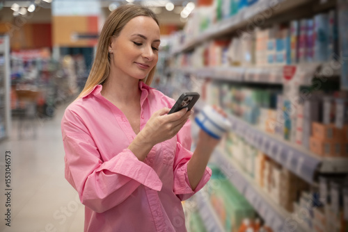 Young woman in a pink shirt selecting healthcare products in a pharmacy, scanning labels with her smartphone while walking through aisles filled with goods