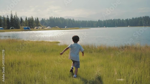 A young boy walks through tall grass near a serene lake. Tents are visible in the background, surrounded by trees. The scene captures a peaceful summer camping experience.