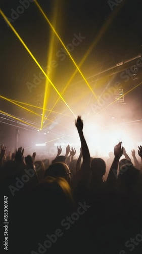A lively concert scene with a diverse crowd of fans raising their hands. Bright yellow laser lights illuminate the atmosphere, creating an energetic vibe.