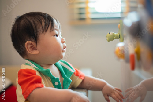 Baby stands up while holding onto playpen wall and looking at the toy stick on the cabinet with natural light