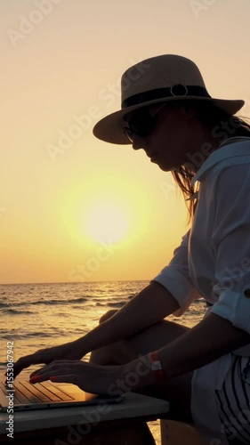 Young woman in sunglasses and sun hat, uses laptop, sitting on the beach by the sea, at sunset or sunrise. freelancer, remote work. work and vacation at sea