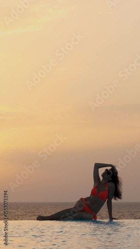 Young woman in swimsuit and sunglasses, is lying and relaxing on edge of outdoor infinity pool with panoramic sea view, at sunrise. travel and vacation concept
