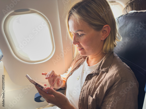 Passenger woman is flying in plane. Girl using phone sitting by airplane window. Traveling female with smartphone inside plane enjoying flight. Woman using wifi connection on board. Traveling girl
