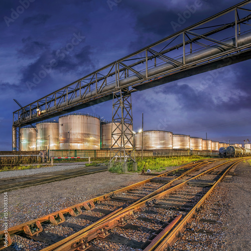 Industrial scene whit rails on the foreground, shiny silos on the background and al large pipeline bridge, Port of Antwerp, Belgium