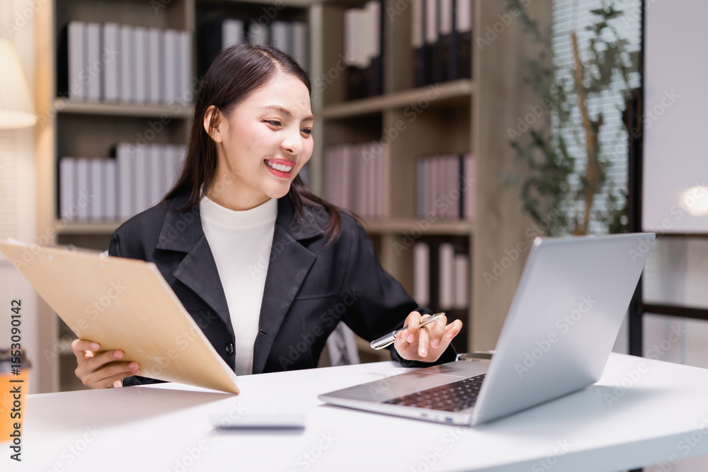 Young businesswoman is smiling while working with a laptop and holding a folder in her office, suggesting a productive and positive work environment Young businesswoman is smiling while working with a laptop and holding a folder in her office, suggesting a productive and positive work environment