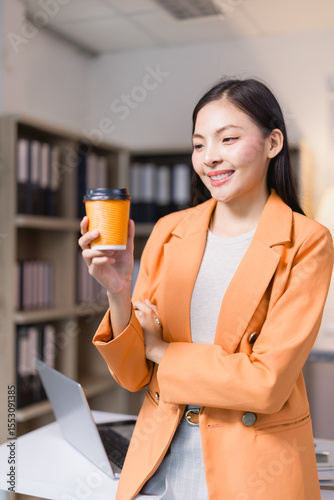 Young asian businesswoman in orange suit standing with arms crossed holding takeaway coffee cup and smiling in modern office with laptop on desk