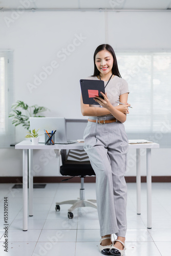 Asian businesswoman leaning on a desk, smiling while using a tablet and holding a pen in her other hand. Modern office setting with natural light enhancing her professional appearance