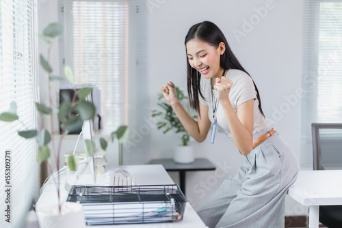 Young Asian businesswoman celebrating success in a modern office, radiating joy and excitement over a recent achievement while working at her desk with a laptop