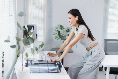Smiling and working at her computer in a modern office, a young businesswoman enjoys a productive workday, embracing the efficiency and creativity of her corporate environment