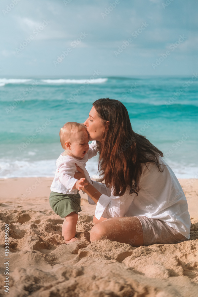 Portrait of mother and baby on vacation on the beach by the ocean. The little boy is capricious and crying, mother calms him down kisses and tickles him to cheer him up. Mothers Day. Travel with child Portrait of mother and baby on vacation on the beach by the ocean. The little boy is capricious and crying, mother calms him down kisses and tickles him to cheer him up. Mothers Day. Travel with child