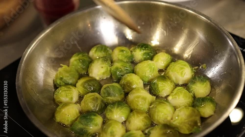 Stirring spoon, kitchen setting, cooking veggies. Close-up captures Brussels sprouts being cooked in stainless pan, stirred by spoon held by hand, showcasing active and healthy cooking fresh produce.