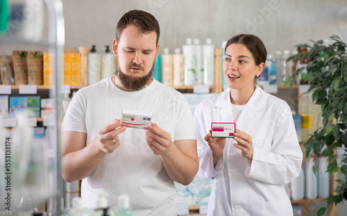 Young man buyer choosing box of paracetamol tablets with help of young woman pharmacist in pharmacy