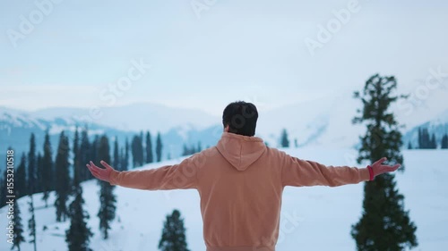 4K Rear view shot of an Indian male tourist raising his arms in front of the snowy Himalayan mountain peaks during the winter season at Gulmarg in Jammu and Kashmir, India. Man enjoys winter season.