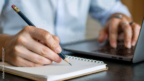 A person taking notes in a notebook with a pencil next to a laptop, indicating study, office work, or creative writing at a desk and reflecting productivity.