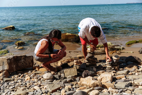 A teenage boy and his mother create stone towers on a sunny beach, surrounded by a rocky shore and calm waves under a bright blue sky.