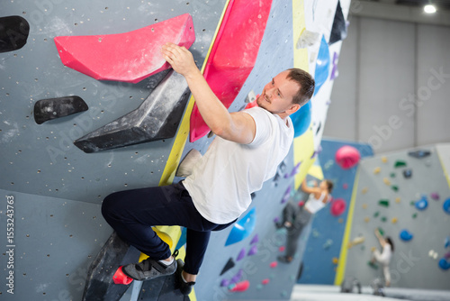 Man climbs steep artificial wall in sports complex and trains endurance and muscle strength. Active recreation in fitness center gym, modern types of recreation photo