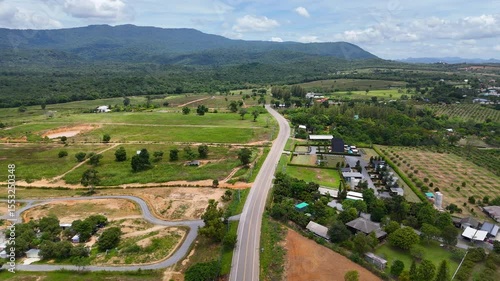 Drone footage the aerial view of a peaceful countryside road winds through green farmland and forest in Wang Nam Khiao, Thailand. A scenic aerial landscape combining rural charm with natural beauty 