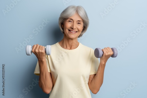 Senior woman with gray hair, smiling while holding light dumbbells in both hands, showcasing fitness and wellness, against a soft blue background, promoting healthy lifestyle choices