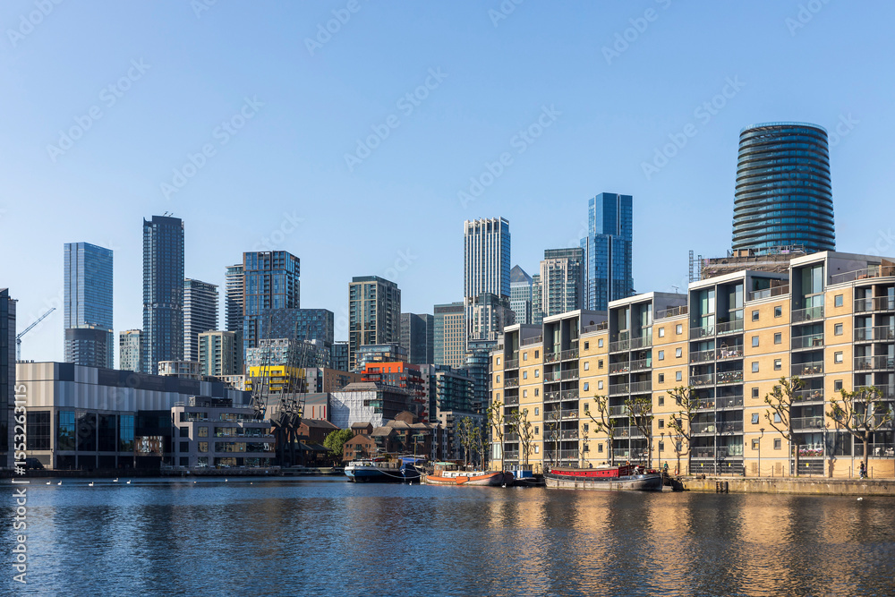 Obraz premium London, UK, 12 May 2025, Canary Wharf A panoramic view of a modern cityscape with numerous tall buildings and a distinctive cylindrical skyscraper, overlooking a wide body of water with moored boats.