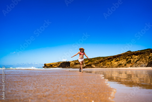 Beautiful mid adult woman walking on sandy beach in summertime. Front view. Monte Clerigo beach on Algarve coast in Portugal