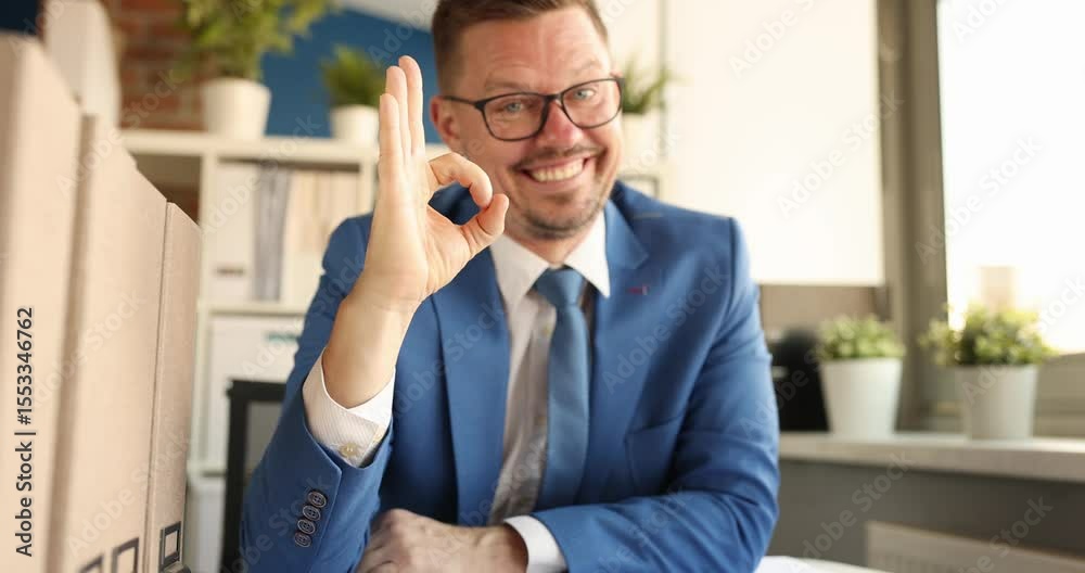 Cheerful businessman in blue suit sits at desk smiling and making OK sign with hand in modern office setting. Man indicates approval and agreement Cheerful businessman in blue suit sits at desk smiling and making OK sign with hand in modern office setting. Man indicates approval and agreement