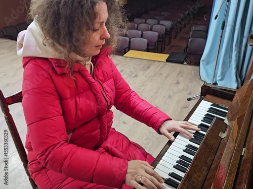 Woman in a red coat is playing the piano. She is wearing a white scarf. The piano is in front of her