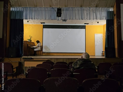 Man sits in a chair in front of a projector screen. A woman is playing the piano in the background in assembly hall