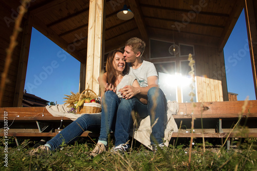 happy pretty couple in jeans and T-shirts is sitting and smiling on the step on the wooden house background on the sunset picnic, young family concept, free space