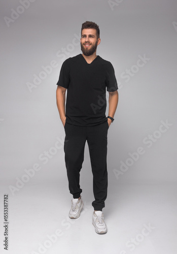 young stylish bearded male nurse with long legs in black medical costume is standing in step with hands in pockets, looking straight and smiling on gray wall background. medical concept. free space