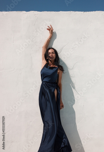 brunette girl in blue long silk dress is standing near white wall background with blue sky at sunny day and smiling cute with hand up in dance. event mood. greek villa. travel concept, free space