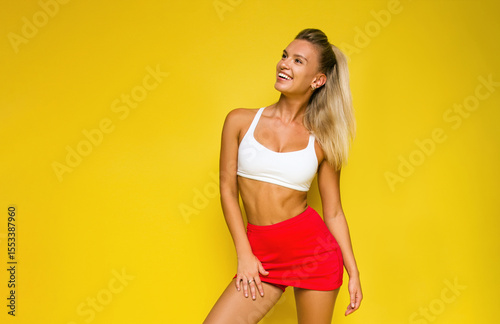 young blonde sports girl in white top and red tennis skirt is standing casual on the yellow wall background and looking up with happy smile. sport lifestyle concept, free space