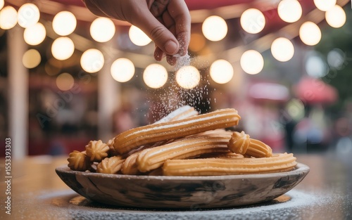 A hand sprinkles powdered sugar on a plate of churros in front of a bokeh background.
