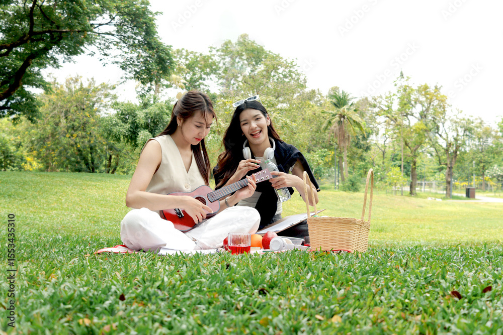 Naklejka premium Happy two beautiful Asian women having a picnic in park. Smiling close friends playing ukulele during picnic outdoor in green garden. People spending time together in natural surroundings.