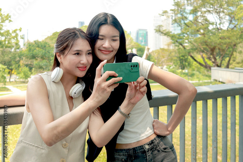 Happy two beautiful Asian women use mobile phone to take a selfie photo together outdoor in park. Smiling close friends hold a book, spend time outside together in green garden. People in nature