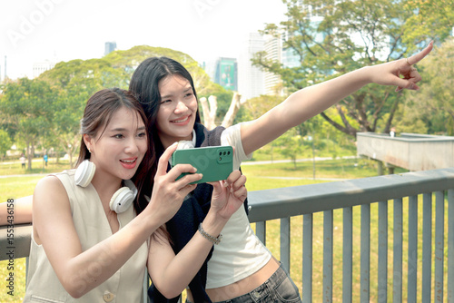 Happy two beautiful Asian women use mobile phone to take a selfie photo together outdoor in park. Smiling close friends hold a book, spend time outside together in green garden. People in nature