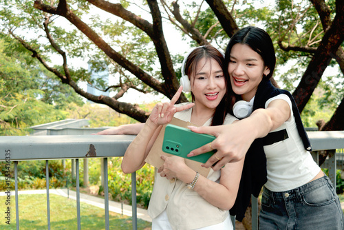 Happy two beautiful Asian women use mobile phone to take a selfie photo together outdoor in park. Smiling close friends hold a book, spend time outside together in green garden. People in nature