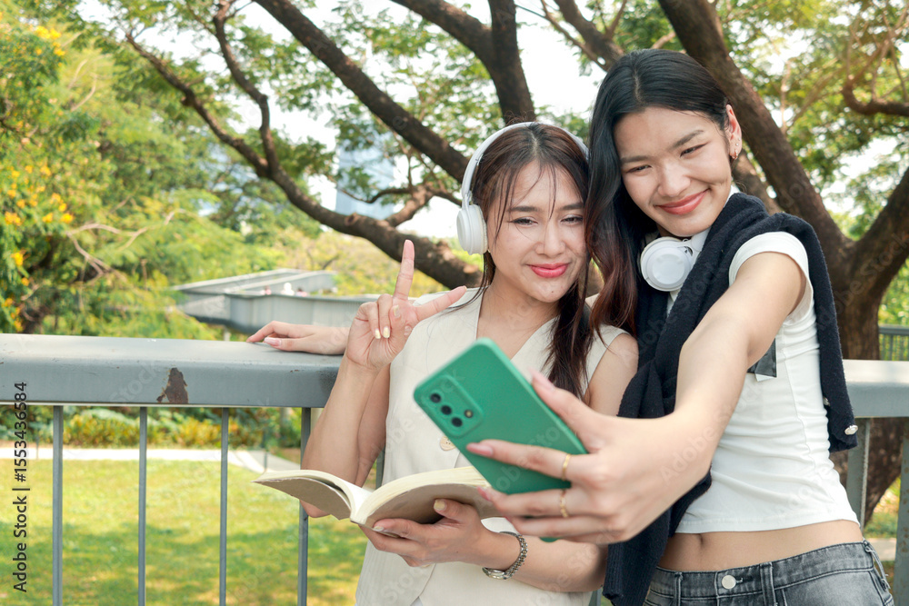 Fototapeta premium Happy two beautiful Asian women use mobile phone to take a selfie photo together outdoor in park. Smiling close friends hold a book, spend time outside together in green garden. People in nature