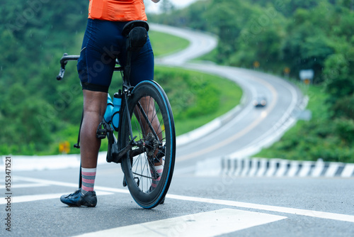 A cyclist in sporty gear pauses on a winding road surrounded by lush green hills.