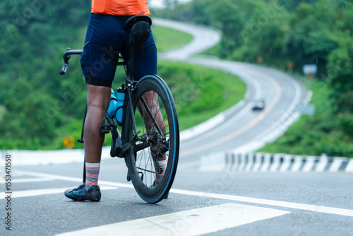 A cyclist in bright gear stands on a paved road with a winding curve ahead, surrounded by green hills and a distant vehicle.