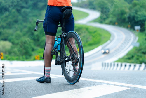 A cyclist wearing colorful socks and an orange jersey pauses with their bike on a wet road near a winding, green hillside.