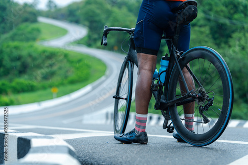 A cyclist with a road bike pauses on a winding road surrounded by greenery, ready for a ride or rest.