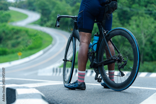 Cyclist standing with a black road bike on a winding road surrounded by green hills and trees.