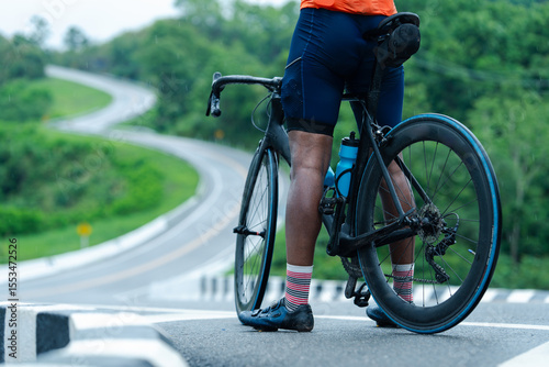 A cyclist in orange gear stands beside a black road bike on a winding country road surrounded by lush greenery.